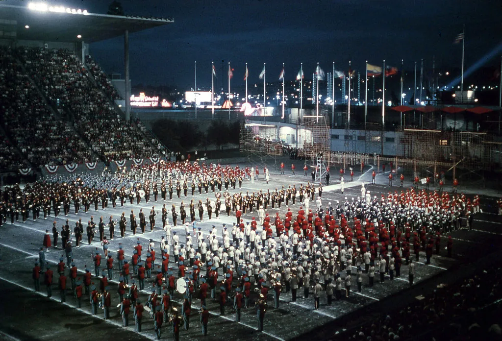 A color photograph of red and white dressed marching bands in the stadium at the Seattle Center at night. The stands are full of people and spotlights hit the band and many nations flags fly in the background.